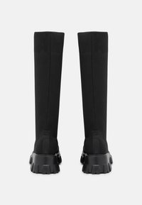 Botas negras hasta la rodilla con parte superior de tejido elástico y suelas de goma gruesas. Detalles acanalados notables en las suelas y acentos de logotipo en los tacones.