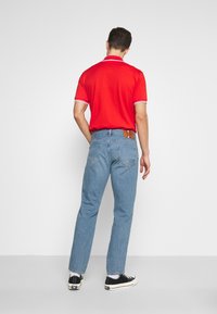 Camisa de polo roja con acentos blancos, combinada con jeans azul claros y zapatillas negras. Los jeans tienen una etiqueta de cuero en la parte posterior.