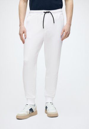 Mand iført hvide joggingbukser med sort snor og hvide sneakers med sorte og beige detaljer, der står mod en ensfarvet baggrund.