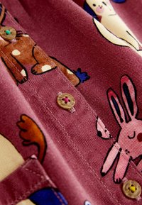 Rosa Cordstoffstoff mit Cartoon-Tiermotiven, darunter ein Kaninchen und ein Hirsch. Ein hellbrauner Knopf mit rotem Fadendetail ist sichtbar.