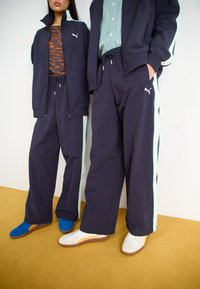 Ναυτικές φόρμες με λευκή και μέντα λωρίδα στο πλάι, συνδυασμένες με πολύχρωπα παπούτσια—μπλε και λευκά με πορτοκαλί λεπτομέρειες—σε κίτρινο πάτωμα.