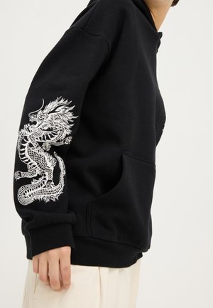 Black hoodie with white detailed dragon graphic on sleeve, worn by person standing against plain background, hand relaxed by side.