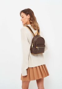 Zaino marrone con motivo e dettagli beige, forma arrotondata, tasca frontale con zip e spallacci regolabili, indossato sulla schiena sopra un maglione chiaro.