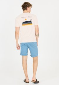 Ljusrosa t-shirt med en California Surf Club grafik, med berg och en sol, ihopparad med rullade blå shorts och bruna sandaler.