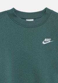 Žalias džemperis su klasikine apvalia iškirpte ir ribbintais rankogaliais, priekyje baltas „Nike“ logotipas. Lygios audinio faktūros.