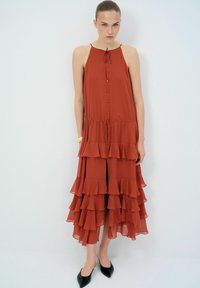 Robe maxi en orange rouille, avec des volants en cascade, un col à nouer, une fermeture à boutons à l'avant et un ourlet fendu. Portée avec des ballerines noires à bout pointu.