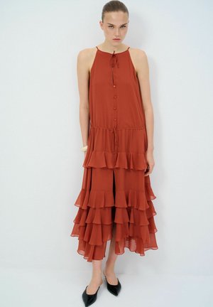 Robe maxi en orange rouille, avec des volants en cascade, un col à nouer, une fermeture à boutons à l'avant et un ourlet fendu. Portée avec des ballerines noires à bout pointu.