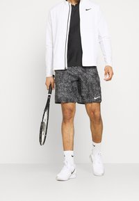 Weiße Sportjacke, schwarzes Polo-Shirt, gemusterte schwarze Shorts, weiße Tennisschuhe, hält einen schwarz-weißen Tennisschläger.