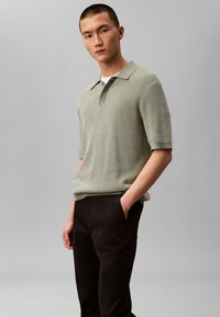 Tricou polo verde deschis, textură tricotată, mâneci scurte, guler clasic. Asortat cu pantaloni negri, mâinile în buzunare, fundal neutru.