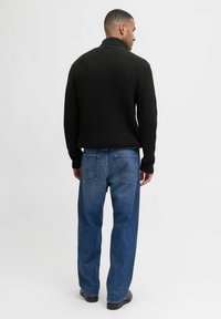 Zwarte gebreide trui met een hoge kraag, gecombineerd met loszittende blauwe denim jeans. Zichtbare stikdetails op de achterzakken.