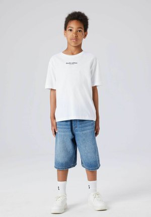 Dječak stoji okrenut prema naprijed, obučen u bijelu majicu s natpisom, traper kratke hlače do koljena, bijele čarape i bijele tenisice na jednobojnoj pozadini.