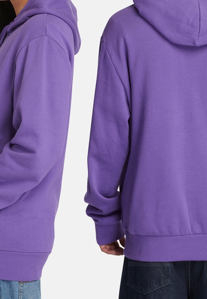 Due persone con felpe viola con cappuccio, viste da metà torace ai fianchi, una rivolta verso sinistra e l'altra di spalle, su uno sfondo bianco uniforme.