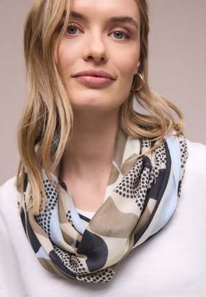 Młoda kobieta o blond włosach, nosząca złote kolczyki koła oraz wzorzysty beżowy, czarny i jasnoniebieski szalik na białej bluzce.