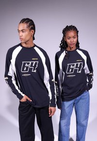 Black long-sleeve shirts with white accents, featuring a bold "64" graphic and text in gold. Loose fit with contrasting sleeve design.