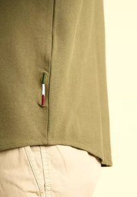 Maglietta a maglia verde oliva con orlo dritto. Presenta un piccolo dettaglio a bandiera multicolore sulla cucitura laterale. Tessuto testurizzato, design informale.