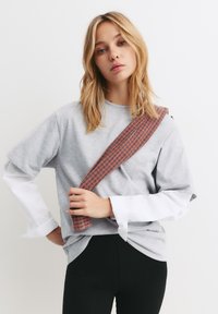 Šedé bavlněné tričko s krátkým rukávem, vrstvené přes bílé tričko, doplněné černými legínami. Obsahuje kostkovaný hnědý popruh přes rameno.