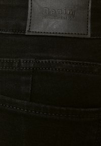 Tela de mezclilla negra con una superficie texturizada. Presenta un parche rectangular de cuero etiquetado como "Denim" con costuras a lo largo de los bordes.