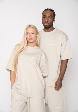 Two models, one female and one male, wearing matching beige oversized t-shirts and shorts with "MULTIPLY" text, standing against a plain background.