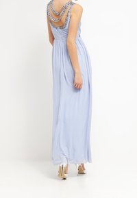 Abito maxi azzurro chiaro con gonna drappeggiata, tre spalline decorative a catena e corpetto aderente. Presenta una texture liscia e una leggera lucentezza.