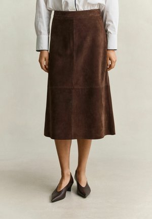 Femme portant une jupe midi en daim marron, une chemise blanche à manches longues et des talons pointus marron foncé, debout sur un sol clair.