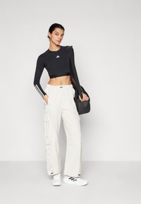 Črn podaljšan crop top z dolgimi rokavi in logotipom, bež cargo hlače in bele superge s črnimi akcenti. Model drži črno športno torbo.