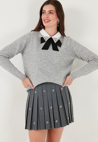 Kobieta w szarym swetrze z ozdobnym białym kołnierzykiem i czarną kokardą, połączonym z szarą plisowaną spódnicą zdobioną diamentowymi akcentami, uśmiechająca się.