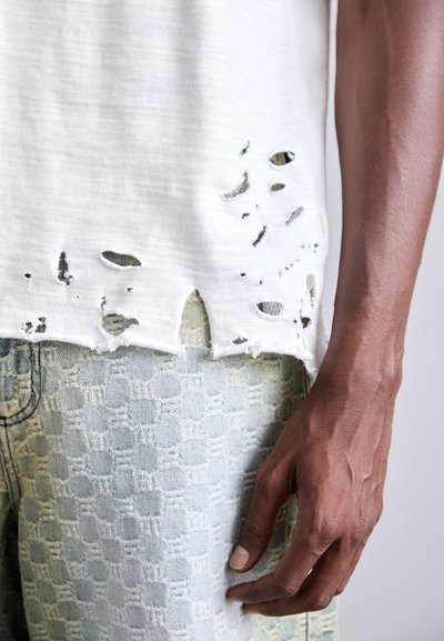 Hvid slidt t-shirt med flere rifter i kanten, fuldendt med lyseblå mønstrede denim shorts. Synlig hånd med definerede årer.