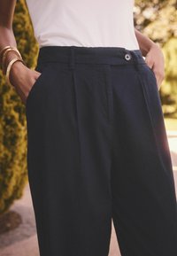 Navyblauwe high-waisted broek met een knoopsluiting, plooien aan de voorkant en zijzakken, gemaakt van een gladde stof.