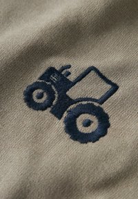 Bēša auduma ar tumši zilu izšūtu traktora dizainu, izceļot apaļus riteņus un taisnstūrveida kravas zonu. Teksturēts un detalizēts šuvums.