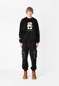 Sudadera negra con estampado gráfico, combinada con pantalones de chándal negros con bolsillos con cremallera y botas marrones. Diseño casual y de corte relajado.