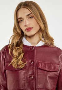 Burgundy nahkake jacket, jossa on kaksi etutaskua, nepparipainikkeet ja valkoinen kaulus. Sileä pinta ja moderni muotoilu.