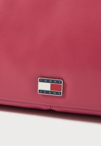 Rosa skinnveske med rektangulær form, glatt tekstur og en fremtredende merketagg med blå, hvite og røde aksenter.