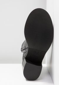 Semelle d'une botte en cuir noir avec un petit talon carré, placée à la verticale contre un mur blanc et une surface blanche.