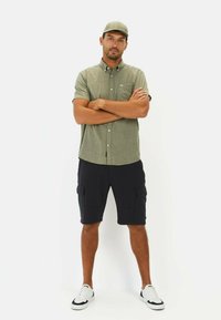 Grøn kortærmet skjorte med knapper, cargo-shorts i sort og hvide sneakers; modellen står med armene cruzet mod en hvid baggrund.