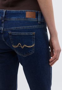 Donkerblauwe spijkerbroek met een bruine leren patch, decoratieve stiksels op het achterzakje en een aansluitend ontwerp met vijf zakken.