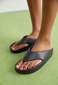 Mustat kumisaappaat, joissa on leveä risteytysremmissuunnittelu. Varusteena teksturoitu pohja ja hillitty Nike-logot sivussa.