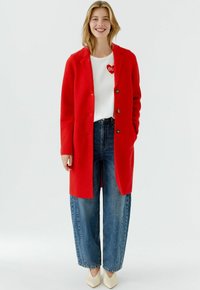 Red wool coat with a notched collar, front buttons, and side pockets, paired with white shirt and wide-leg blue jeans. Cream-colored shoes.
