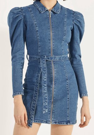 Kvinde iført en blå denim minikjole med lange ærmer, puffede skuldre, en lynlås foran, krave og et matchende stofbælte.