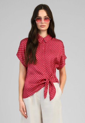 Femme portant un chemisier rouge à manches courtes à pois avec un nœud sur le côté et un pantalon blanc, posant avec une main dans la poche et portant des lunettes de soleil roses.