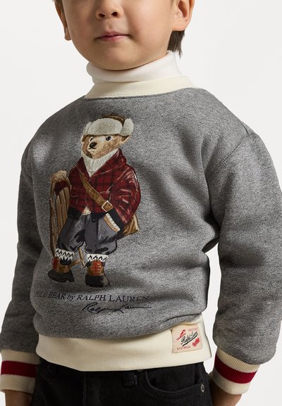 Sudadera gris con un oso estampado que lleva una chaqueta de cuadros rojos, pantalones de nieve y un trineo. Cuenta con puños acanalados con detalles en rojo y crema. Etiqueta visible.