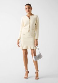 Tenue couleur crème comprenant une veste ajustée texturée ornée d'embellissements et une jupe assortie avec un ourlet à volants. Accessoirisée d'un sac à main argenté.