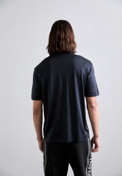 Persoană cu păr brunet până la umeri, purtând un tricou simplu negru cu mâneci scurte și pantaloni negri cu dungi verticale albe.