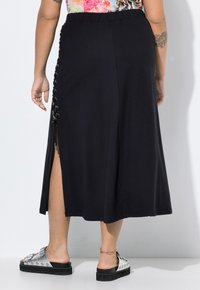 Schwarze, knielange Rock mit seitlichem Schnürdetail und Reißverschlussakzent. Glattes Gewebe mit fließender Silhouette. Kombiniert mit silbernen Sandalen.