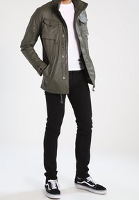 Man wearing olive green zip-up jacket with pockets, white t-shirt, black pants, and black and white sneakers, standing with one hand in pocket.