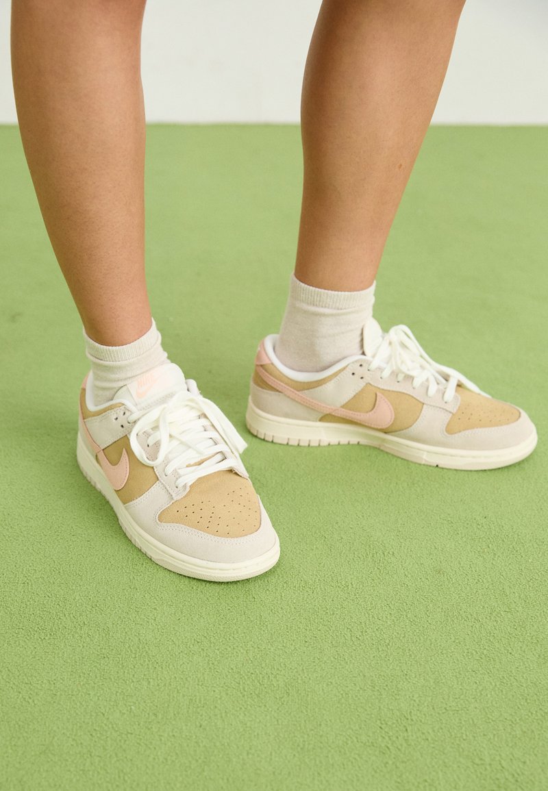 Beżowe i białe sneakersy z różowymi akcentami, z cholewką wykonaną z skóry i zamszu, perforowanym przodem oraz płaskimi sznurowadłami, umieszczone na zielonym tle.