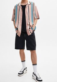 Camicetta a maniche corte con bottoni, a strisce rosa, teal e crema, indossata sopra una maglietta nera. Abbinata a pantaloni corti neri e scarpe da ginnastica.