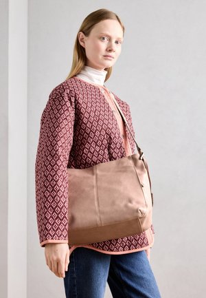 Brun læderhandtaske med en glat overflade, der har to udvendige sømme, en justerbar rem og bæres med en mønstret maroon jakke.