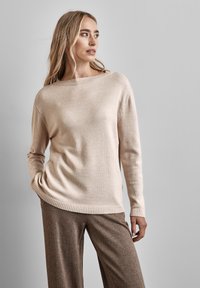 Beige stickad tröja med bred båturringning, ribbade muddar och fåll. Kombineras med texturerade bruna vida byxor. Slät tygtextur.