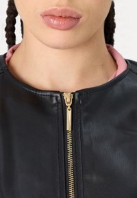 Primo piano delle labbra e del collo di una persona che indossa una giacca di pelle nera con una zip dorata parzialmente chiusa su un collare rosa.