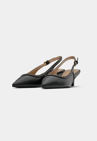 Escarpins slingback noirs en cuir à bout pointu avec de petits clous le long des bretelles et talons petits sur fond blanc.